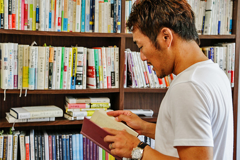 本のぎっしり詰まった本棚の前で1冊を手に取る濱本さん